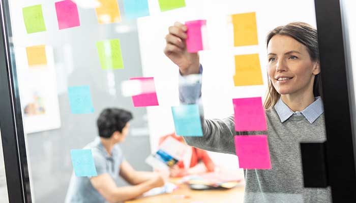 Nainen kiinnittää värikkäitä post-it lappuja ikkunaan, taustalla mies ja nainen sekä värillisiä asiakirjoja